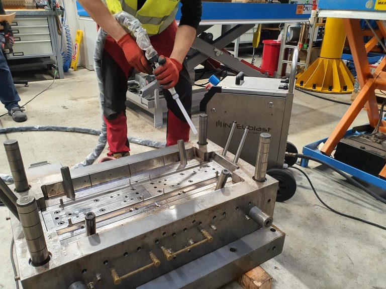 4 godziny zamiast 2 dni. Jak czyszczenie suchym lodem uratowało weekendową produkcję 4 Osoba ubrana w czerwone spodnie, rękawice i żółtą kamizelkę ochronną czyści dużą metalową formę za pomocą śrutownicy w warsztacie przemysłowym. W tle widoczne są różne urządzenia i narzędzia.