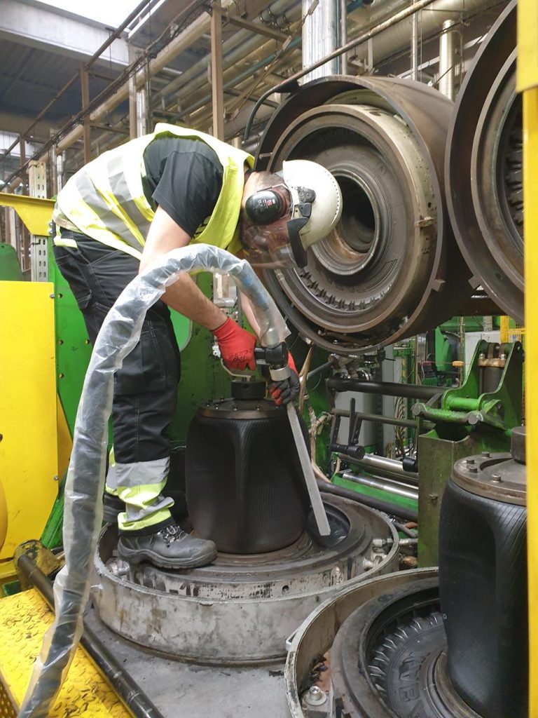 Wynajem maszyny do czyszczenia suchym lodem – kiedy to się opłaca? 3 Pracownik noszący sprzęt ochronny i kamizelkę odblaskową obsługuje maszyny w środowisku przemysłowym, używając węża do napełniania lub czyszczenia dużego elementu silnika otoczonego rurami i metalowymi konstrukcjami.