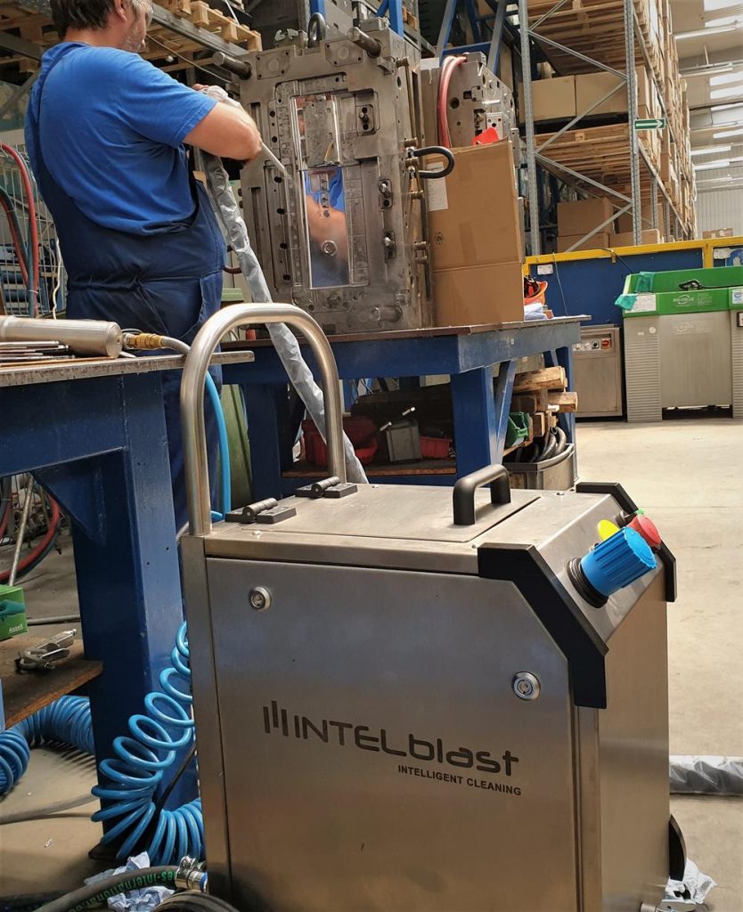 Jak bezpiecznie obsługiwać maszynę do czyszczenia suchym lodem? Poradnik 1 Osoba pracuje w warsztacie przy użyciu dużej maszyny, podczas gdy na pierwszym planie znajduje się metalowe urządzenie czyszczące INTELblast. W tle widoczne są półki i różne urządzenia.