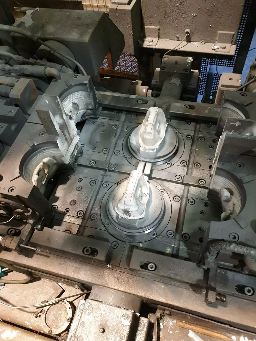 Przemysł odlewniczy 2 Zbliżenie formy przemysłowej z dwoma częściowo uformowanymi białymi odlewami wewnątrz, otoczonej metalowymi maszynami i pyłem, podkreśla znaczenie Czyszczenia Form Odlewniczych Suchym Lodem w środowisku produkcyjnym lub odlewniczym.