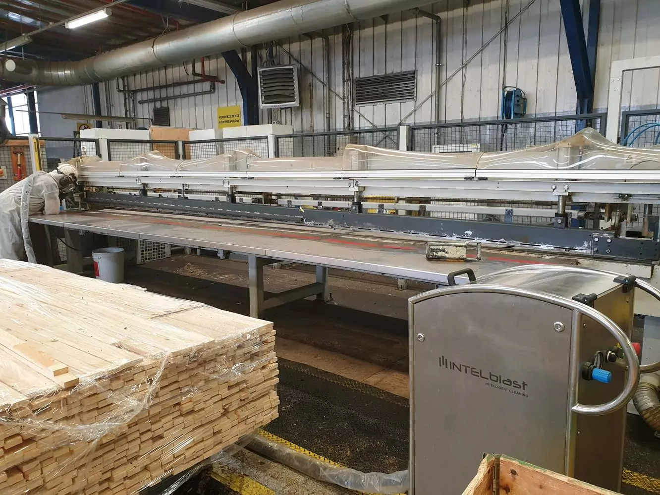 Przemysł drzewny 4 Duża maszyna przemysłowa z napisem INTELblast jest ustawiona w fabryce Czyszczenia Maszyn Stolarskich Suchym Lodem, ze stosami drewnianych desek na pierwszym planie i różnymi metalowymi rurami, otworami wentylacyjnymi i sprzętem w tle.
