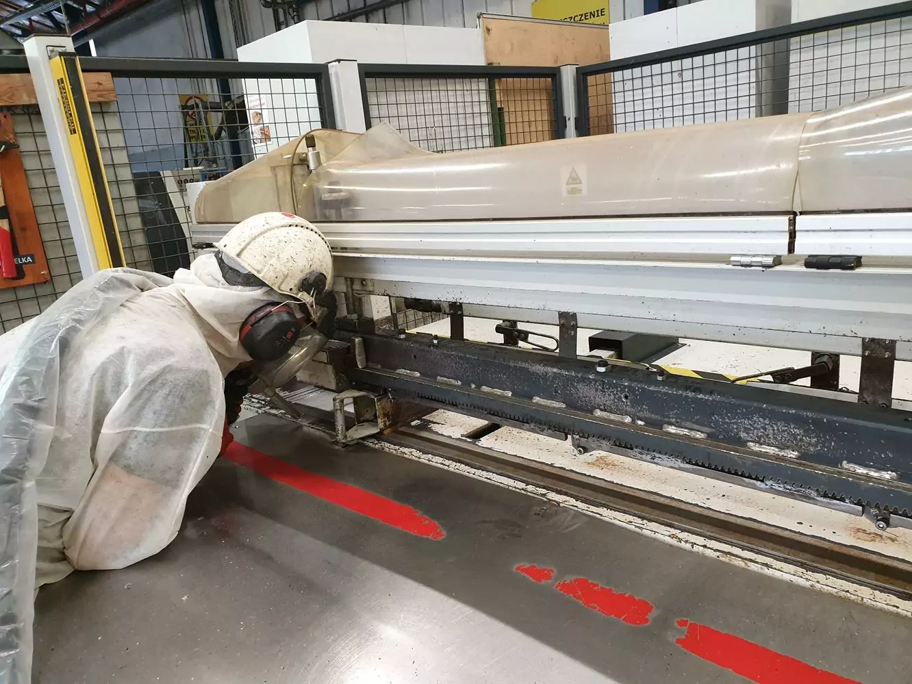 Przemysł drzewny 3 Pracownik w odzieży ochronnej, w tym w kasku i osłonie twarzy, sprawdza maszyny w zakładzie przemysłowym. Czysty, zorganizowany obszar, wyposażony w bariery ochronne, sugeruje, że Czyszczenie Maszyn Stolarskich Suchym Lodem jest wykorzystywane do konserwacji.