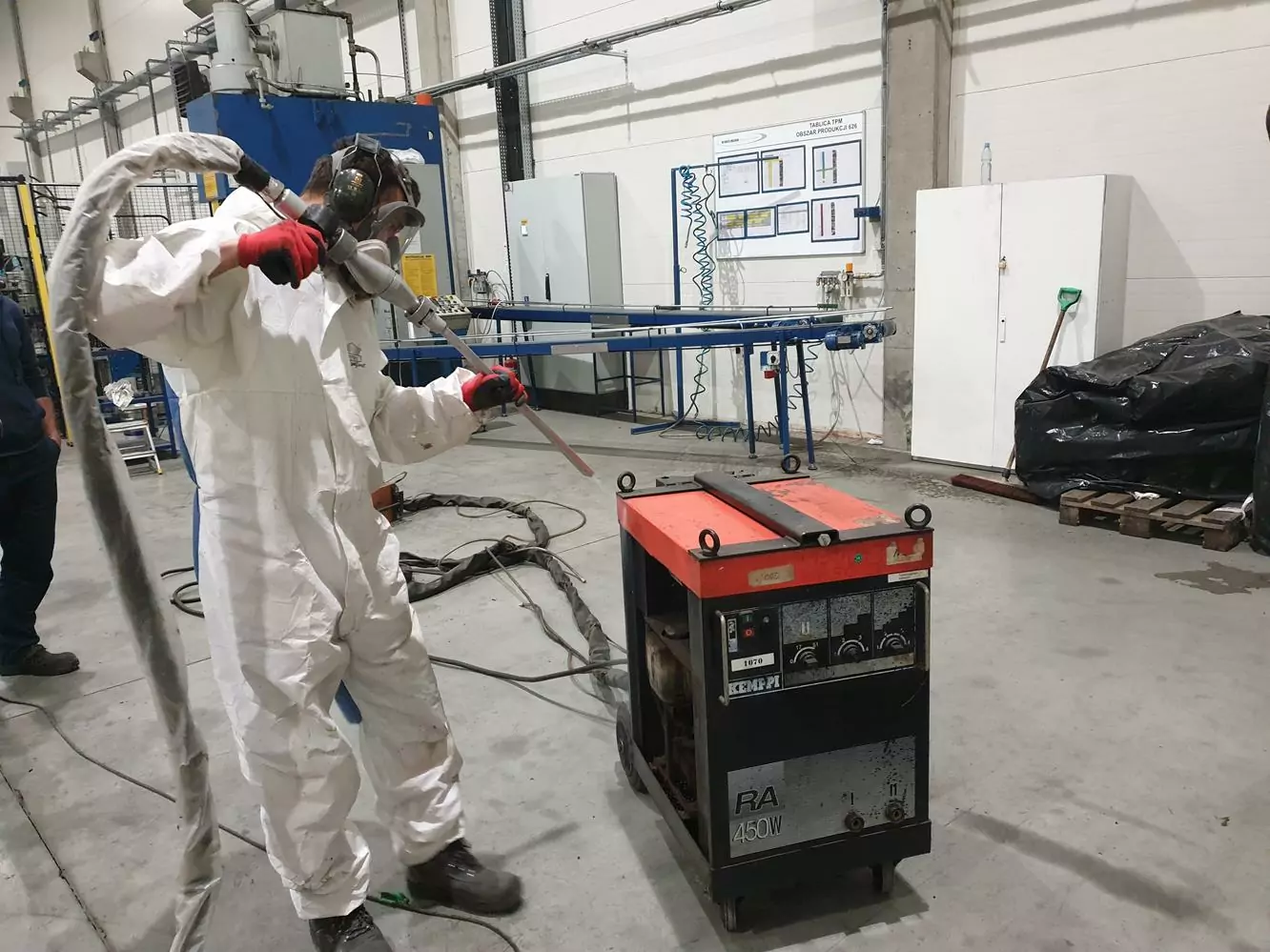 Czyszczenie spawalni i spawarek 4 Osoba w kombinezonie ochronnym i masce obsługuje sprzęt przemysłowy wewnątrz fabryki, trzymając wąż i dyszę skierowaną na czerwono-czarną maszynę na kołach - część Galerii Zdjęć Czyszczenia Suchym Lodem. Z tyłu widoczne są panele sterowania i maszyny.