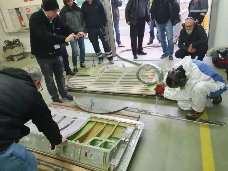 Czyszczenie Suchym Lodem vs. Piaskowanie 7 Grupa ludzi obserwuje, jak dwie osoby badają i pracują nad dużymi, zdemontowanymi metalowymi panelami w warsztacie, porównując Czyszczenie suchym lodem vs piaskowanie. Jedna osoba nosi kombinezon ochronny i maskę, obsługując sprzęt.