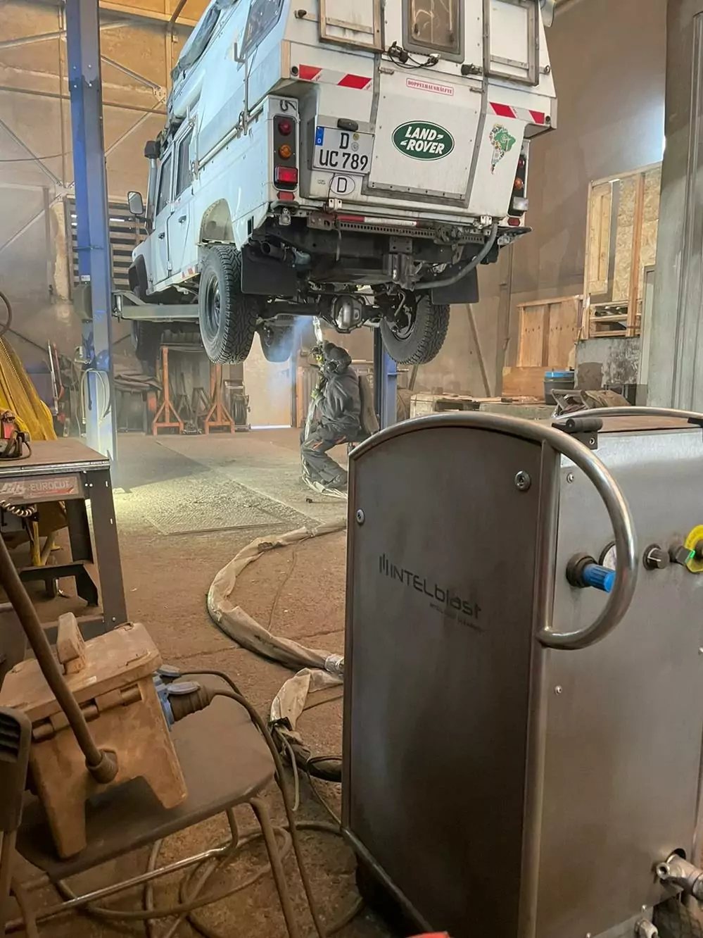 Land Rover jest podnoszony na podnośniku warsztatowym, podczas gdy mechanik pracuje pod nim. Na pierwszym planie duża maszyna Intelblast jest połączona wężami, pokazując proces dla Galerii Zdjęć Czyszczenia Suchym Lodem w ruchliwym warsztacie wypełnionym narzędziami i sprzętem.
