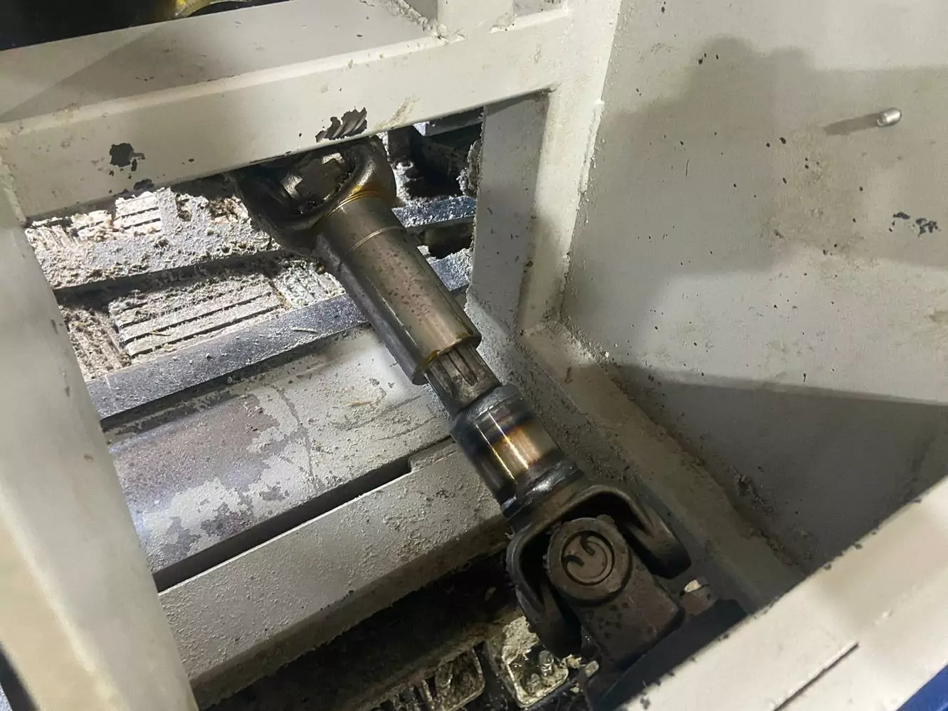 Przemysł drzewny 5 Zbliżenie na metalowy wał przegubu uniwersalnego w maszynie przemysłowej, otoczony białą metalową ramą i zanieczyszczeniami. Widoczne zużycie i brud sugerują potrzebę Czyszczenia Maszyn Stolarskich Suchym Lodem w celu optymalnej konserwacji.