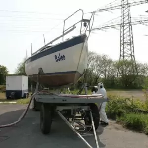 Czyszczenie jachtów i łodzi 2 Osoba w odzieży ochronnej wykonuje Czyszczenie Kadłubów Jachtów Suchym Lodem na kadłubie żaglówki o nazwie Baba, umieszczonej na przyczepie z dużymi liniami energetycznymi i drzewami w tle.