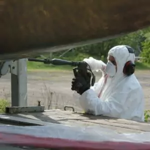 Czyszczenie jachtów i łodzi 3 Osoba w odzieży ochronnej, masce na twarz, rękawicach i nausznikach korzysta z elektronarzędzia na zewnątrz, prawdopodobnie wykonując Czyszczenie Kadłubów Jachtów Suchym Lodem na spodzie dużego obiektu, zapewniając dokładne czyszczenie lub konserwację.