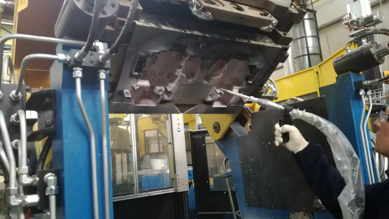 Przemysł odlewniczy 3 Pracownik w rękawicach używa pistoletu natryskowego do powlekania dużej metalowej formy, demonstrując techniki takie jak Czyszczenie Form Odlewniczych Suchym Lodem, wewnątrz obiektu przemysłowego z maszynami i barierami bezpieczeństwa w tle.