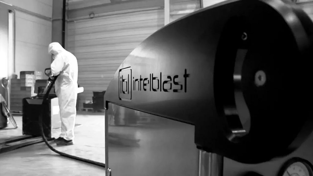 Jaką maszynę do czyszczenia suchym lodem kupić? 6 Osoba w odzieży ochronnej obsługuje sprzęt przemysłowy w dużym obiekcie. Na pierwszym planie maszyna o nazwie Intelblast sugeruje doświadczenie w czyszczeniu przemysłowym - idealna dla tych, którzy zastanawiają się: "Jak wybrać maszynę do czyszczenia takim lodem?.