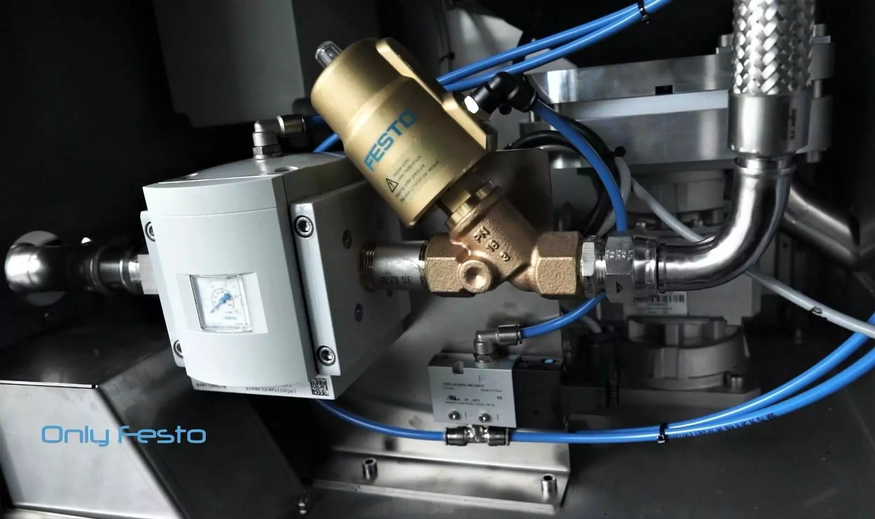 IBL2500 3 Zbliżenie na maszynę przemysłową z zaworem marki Festo z niebieskimi przewodami pneumatycznymi, rurami ze stali nierdzewnej i elementami mechanicznymi. W lewym dolnym rogu widoczne jest logo Festo.