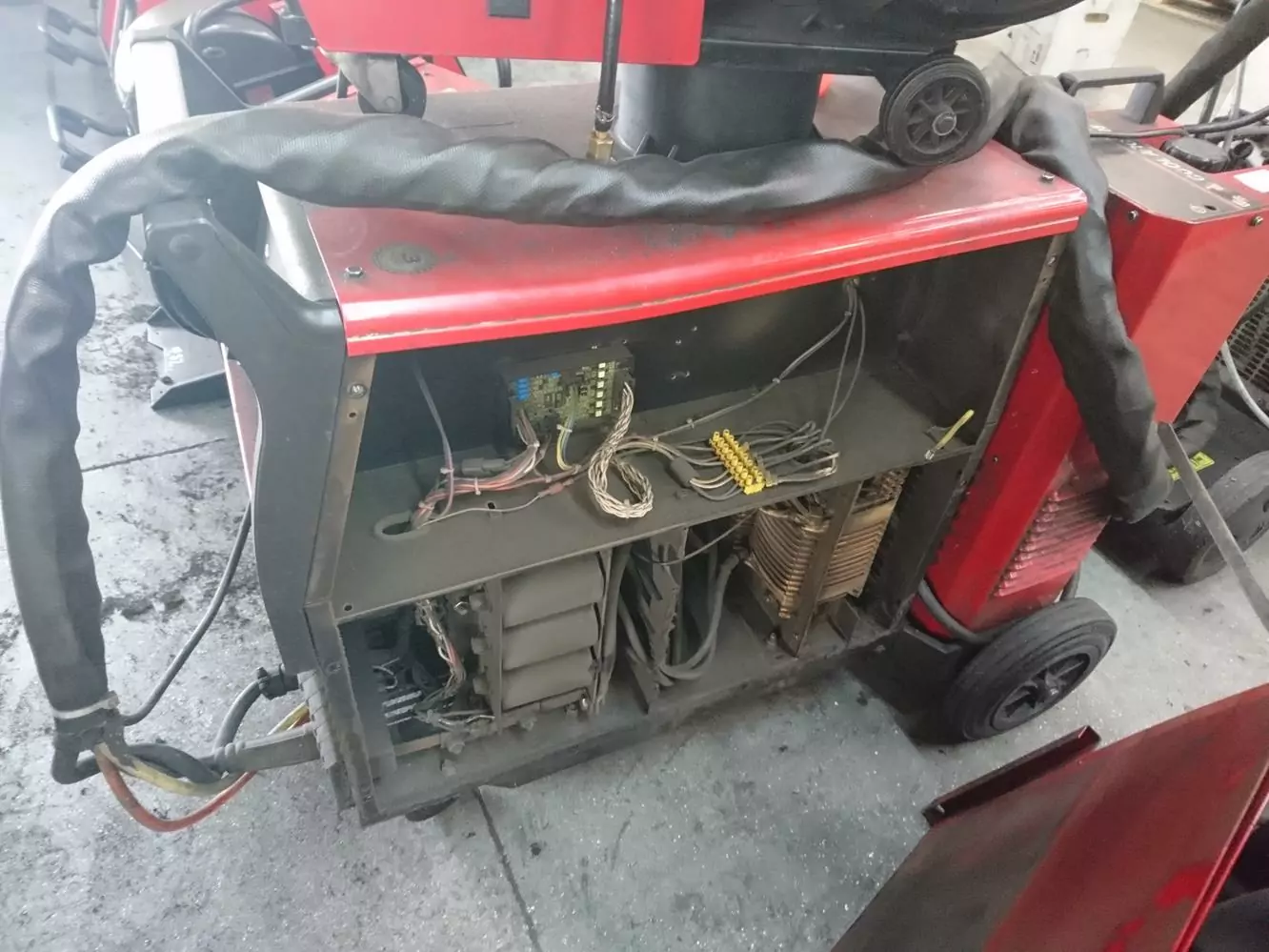 Czyszczenie spawalni i spawarek 2 Czerwona maszyna przemysłowa z otwartym panelem bocznym odsłania wewnętrzne komponenty elektryczne i okablowanie na podłodze warsztatu w Galerii Zdjęć Czyszczenia Suchym Lodem, otoczona narzędziami i sprzętem.