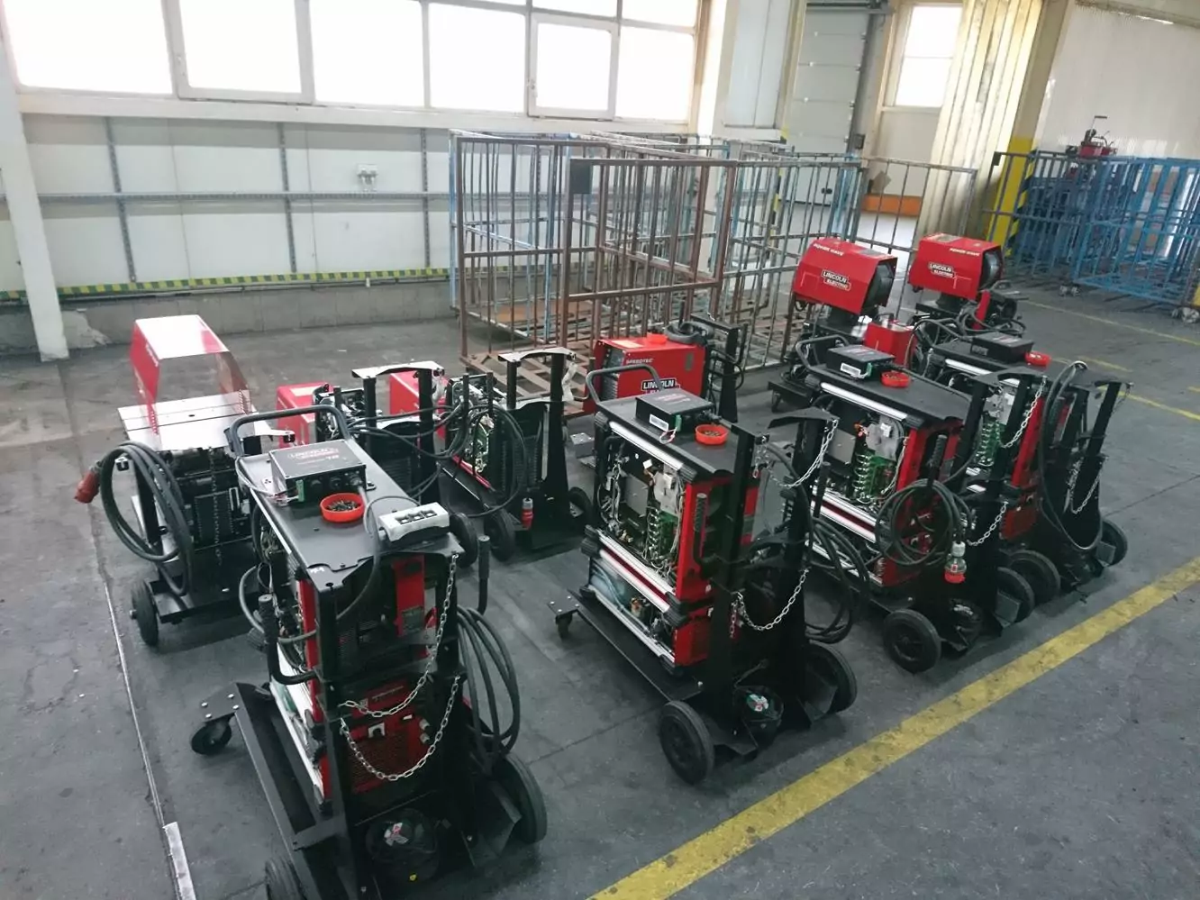 Czyszczenie spawalni i spawarek 5 Na podłodze przestronnego, dobrze oświetlonego warsztatu w Galerii Zdjęć Czyszczenia Suchym Lodem, z dużymi oknami i metalowymi regałami w tle, ustawiono kilka przemysłowych spawarek w czerwono-czarnych obudowach.