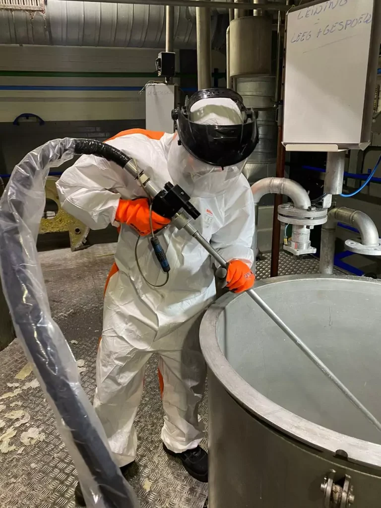 Osoba w stroju ochronnym i kasku używa dużego węża i metalowego pręta, aby wykonać Czyszczenie Maszyn Spożywczych Suchym Lodem na urządzeniach przemysłowych w fabryce lub zakładzie przetwórczym.