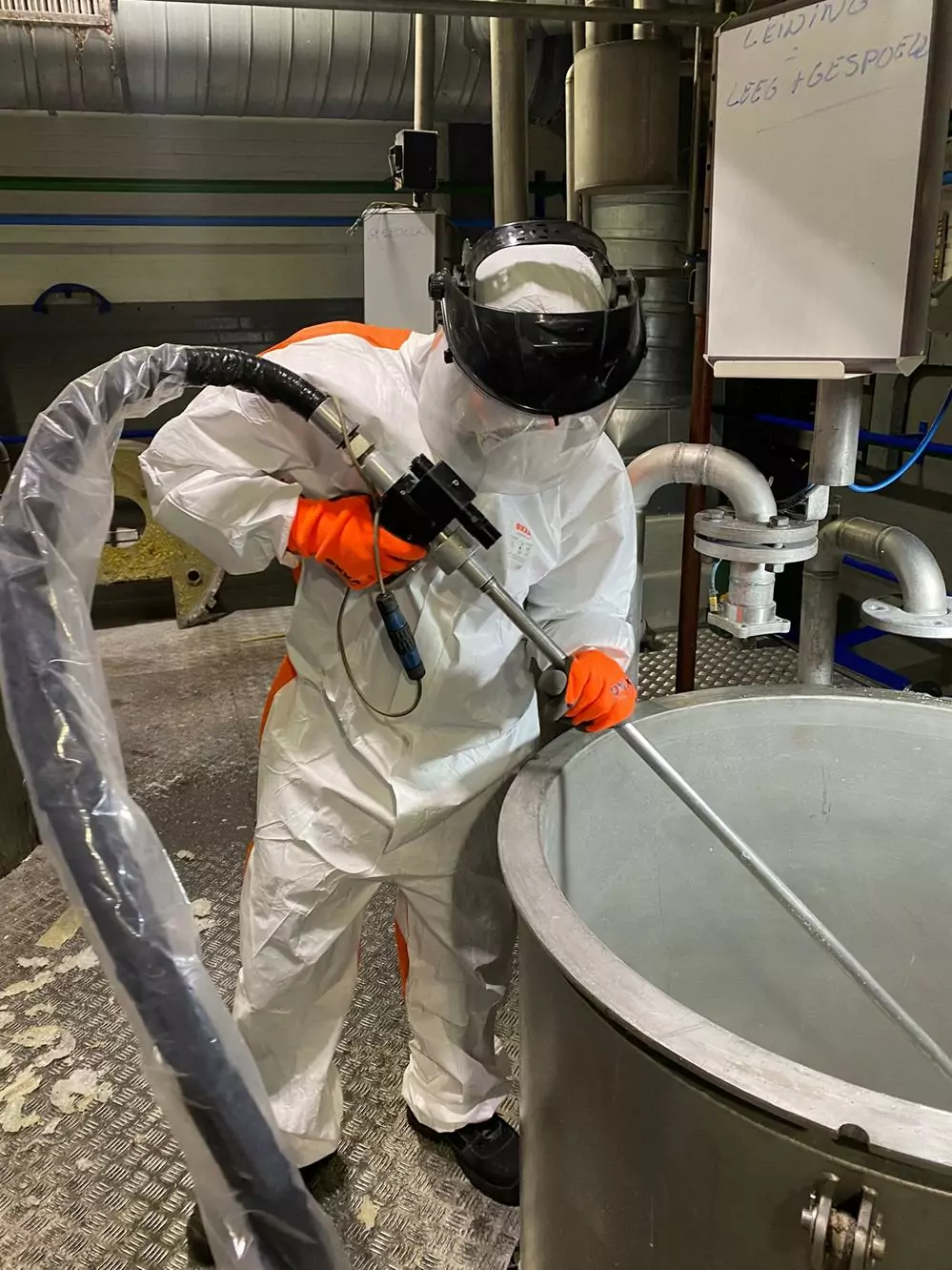 Przemysł spożywczy 2 Osoba w stroju ochronnym i kasku używa dużego węża i metalowego pręta, aby wykonać Czyszczenie Maszyn Spożywczych Suchym Lodem na urządzeniach przemysłowych w fabryce lub zakładzie przetwórczym.