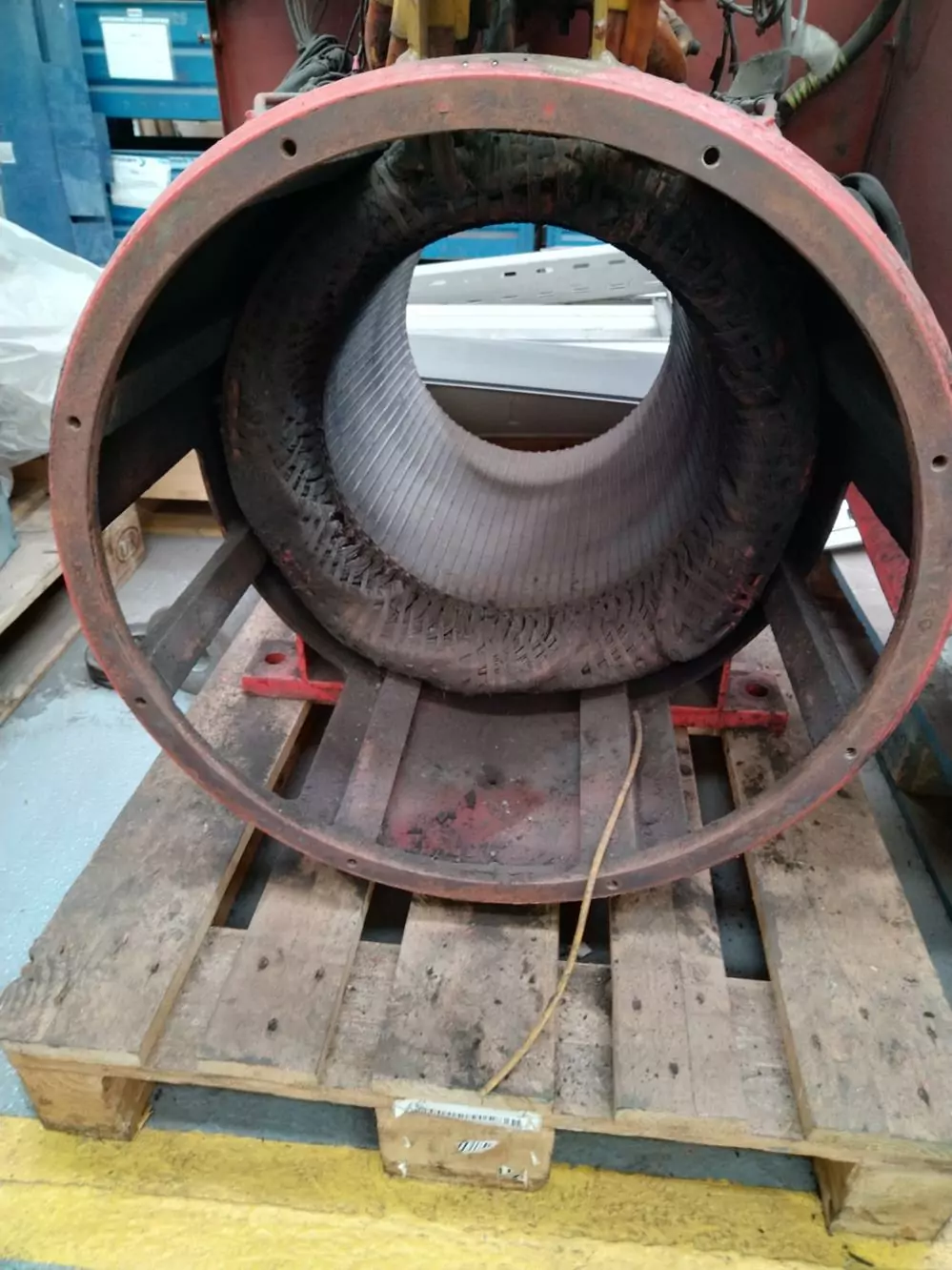 Czyszczenie instalacji i urządzeń elektrycznych 10 Duży wydrążony cylindryczny metalowy element, prawdopodobnie stojan silnika elektrycznego, leży na drewnianej palecie w otoczeniu przemysłowym. Wewnątrz widoczne są rowki i ślady uzwojenia - idealne dla Czyszczenia Rozdzielni Elektrycznych Suchym Lodem.