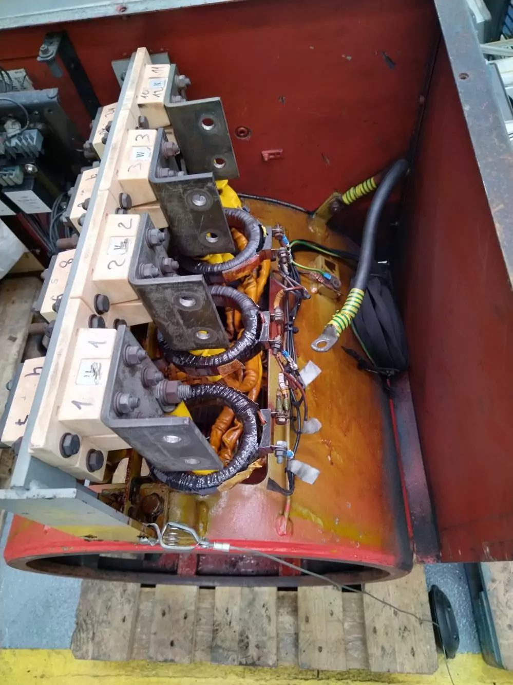 Czyszczenie instalacji i urządzeń elektrycznych 11 Zbliżenie przemysłowej maszyny elektrycznej z miedzianymi cewkami, oznaczonymi zaciskami, przewodami i złączami wewnątrz metalowej obudowy - idealnej do Czyszczenia Rozdzielni Elektrycznych Suchym Lodem, z drewnianymi deskami i żółtym paskiem na podłodze w pobliżu.