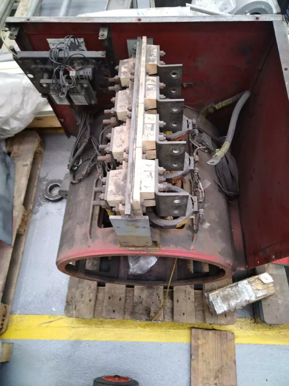 Czyszczenie instalacji i urządzeń elektrycznych 13 Przemysłowy komponent elektryczny z odsłoniętym okablowaniem i zaciskami znajduje się na drewnianej palecie. Jednostka, idealna dla Czyszczenia Rozdzielni Elektrycznych Suchym Lodem, jest częściowo umieszczona w czerwonej metalowej obudowie w ruchliwym warsztacie.