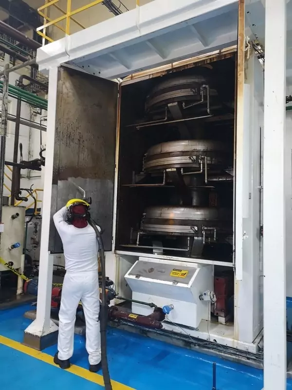 Przemysł spożywczy 3 Pracownik w odzieży ochronnej i kasku stoi przed dużym urządzeniem przemysłowym z trzema okrągłymi metalowymi komorami, które wydaje się wykonywać Czyszczenie Maszyn Spożywczych Suchym Lodem w otoczeniu fabryki.