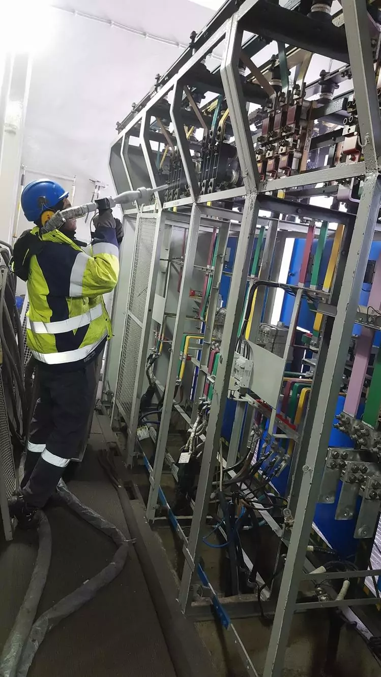 Czyszczenie instalacji i urządzeń elektrycznych 6 Pracownik w kamizelce odblaskowej i kasku kontroluje lub naprawia sprzęt elektryczny, prawdopodobnie wykonując Czyszczenie Rozdzielni Elektrycznych Suchym Lodem, otoczony kolorowymi kablami i metalową ramą w zakładzie przemysłowym.