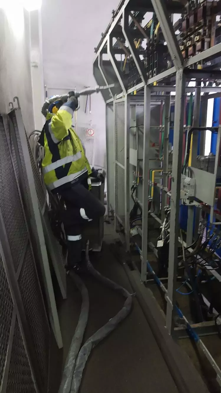 Czyszczenie instalacji i urządzeń elektrycznych 7 Pracownik w kamizelce odblaskowej i sprzęcie ochronnym używa węża do Czyszczenia Rozdzielni Elektrycznych Suchym Lodem, serwisując sprzęt elektryczny w wąskim obszarze przemysłowym wypełnionym kablami i metalowymi konstrukcjami.