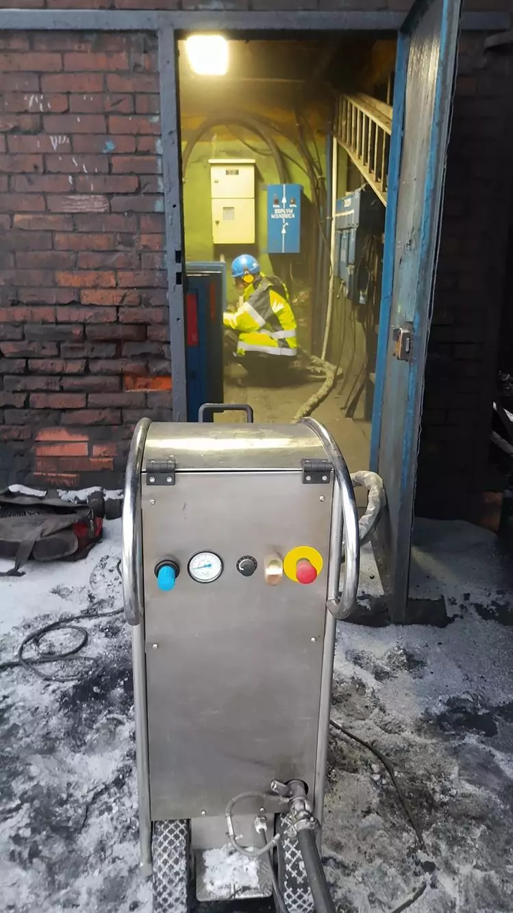 Czyszczenie instalacji i urządzeń elektrycznych 8 Pracownik w ubraniu ochronnym przykuca przy panelach kontrolnych wewnątrz murowanego pomieszczenia przemysłowego, przygotowując się do Czyszczenia Rozdzielni Elektrycznych Suchym Lodem. Na pierwszym planie, za otwartymi niebieskimi drzwiami stoi metalowa maszyna ze wskaźnikami.