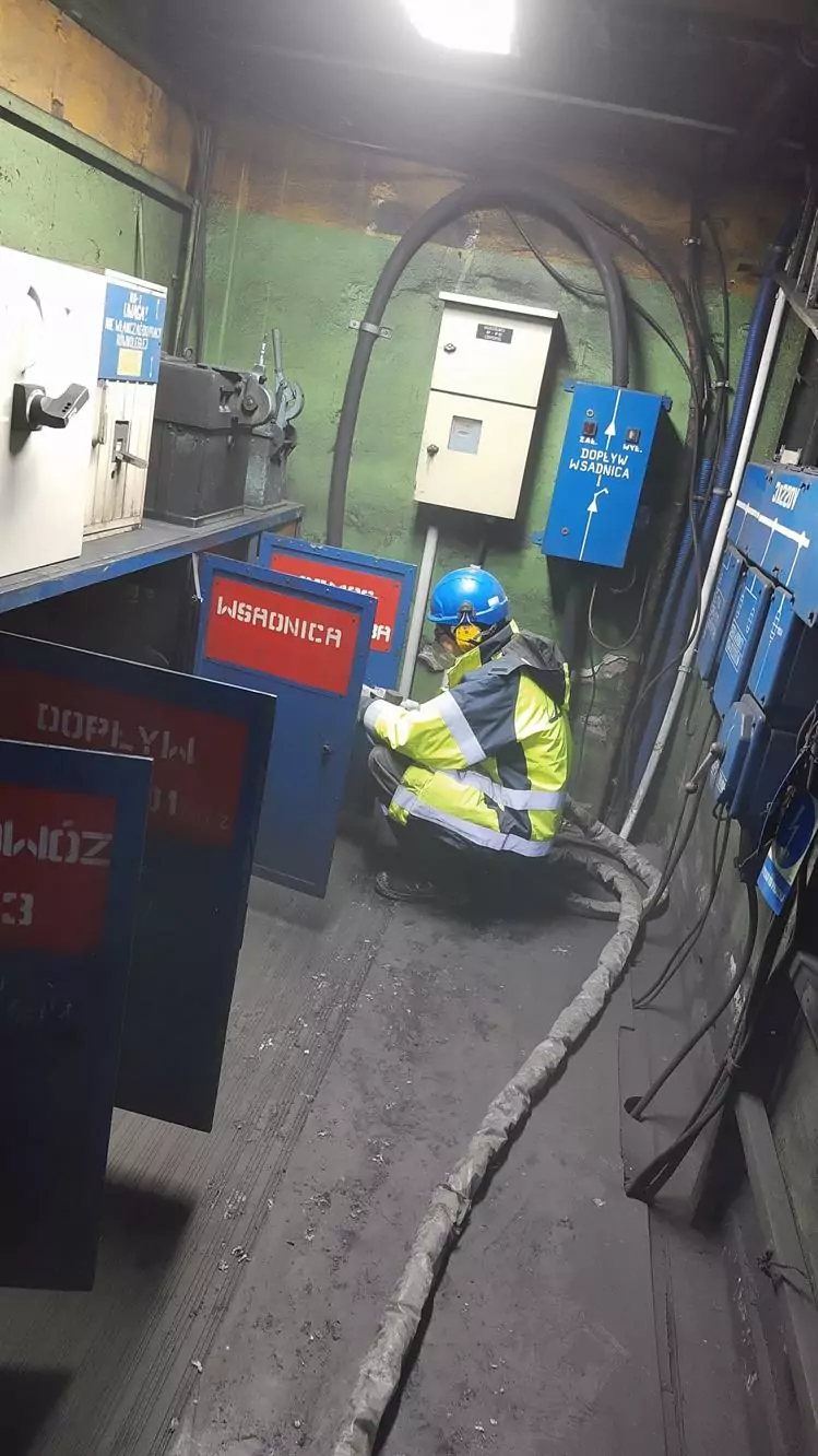 Czyszczenie instalacji i urządzeń elektrycznych 9 Pracownik w dobrze widocznym ubraniu i niebieskim kasku sprawdza sprzęt w pobliżu paneli sterowania, z grubymi kablami na podłodze i niebieskimi skrzynkami elektrycznymi na ścianach - idealne warunki dla Czyszczenia Rozdzielni Elektrycznych Suchym Lodem.