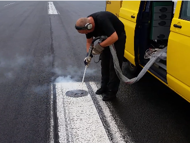 Osoba w odzieży ochronnej używa maszyn do pracy na pasie startowym, tworząc dym podobny do tego w filmie Filmy z Czyszczenia Suchym Lodem. Są obok żółtej furgonetki ze sprzętem do cięcia lub wiercenia w asfalcie.