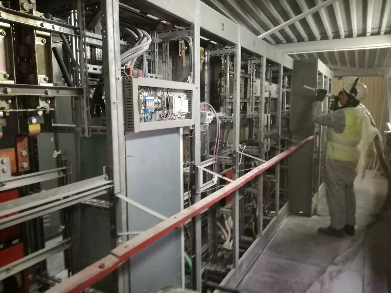 Czyszczenie instalacji i urządzeń elektrycznych 2 Osoba ubrana w sprzęt ochronny i kamizelkę odblaskową sprawdza duży przemysłowy panel sterowania elektrycznego przed wykonaniem Czyszczenia Rozdzielni Elektrycznych Suchym Lodem. Wzdłuż przejścia wewnątrz metalowej konstrukcji biegnie biało-czerwona taśma odgradzająca.