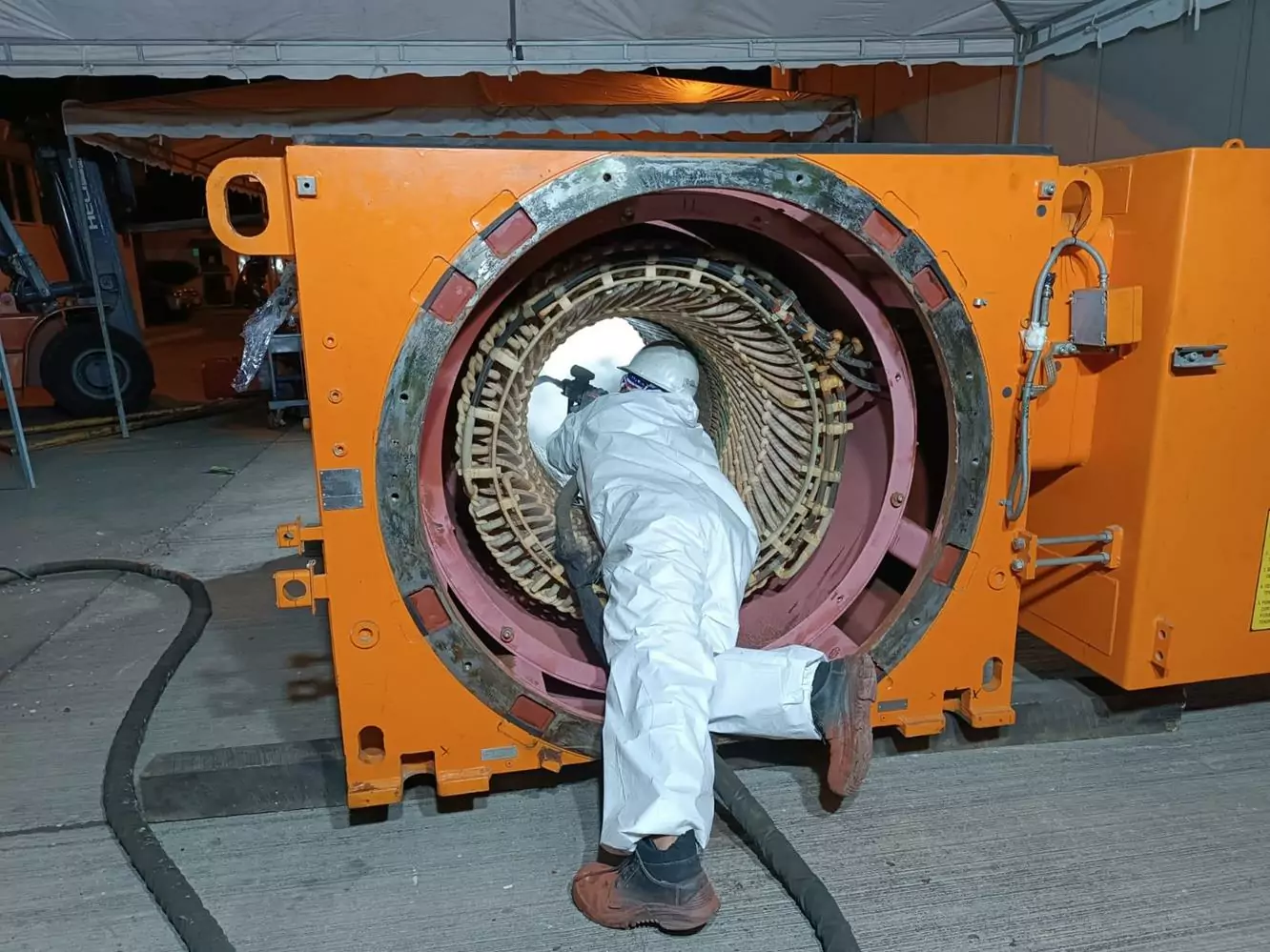 Czyszczenie instalacji i urządzeń elektrycznych 14 Osoba w białym kombinezonie ochronnym i kasku wykonuje Czyszczenie Rozdzielni Elektrycznych Suchym Lodem wewnątrz dużej, cylindrycznej maszyny przemysłowej z pomarańczową obudową, widziana od tyłu pod zadaszeniem na zewnątrz.