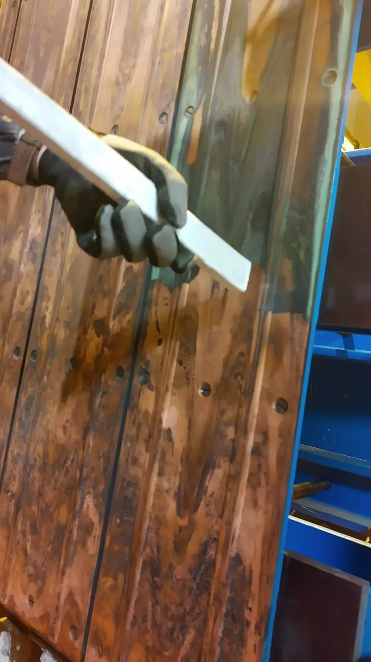 Czyszczenie instalacji i urządzeń elektrycznych 15 Ręka w rękawiczce trzyma białe narzędzie na poplamionej drewnianej powierzchni, prawdopodobnie nakładając lub usuwając powłokę. Niebieskie i żółte obiekty znajdują się w tle, przywołując na myśl precyzję Czyszczenia Rozdzielni Elektrycznych Suchym Lodem.