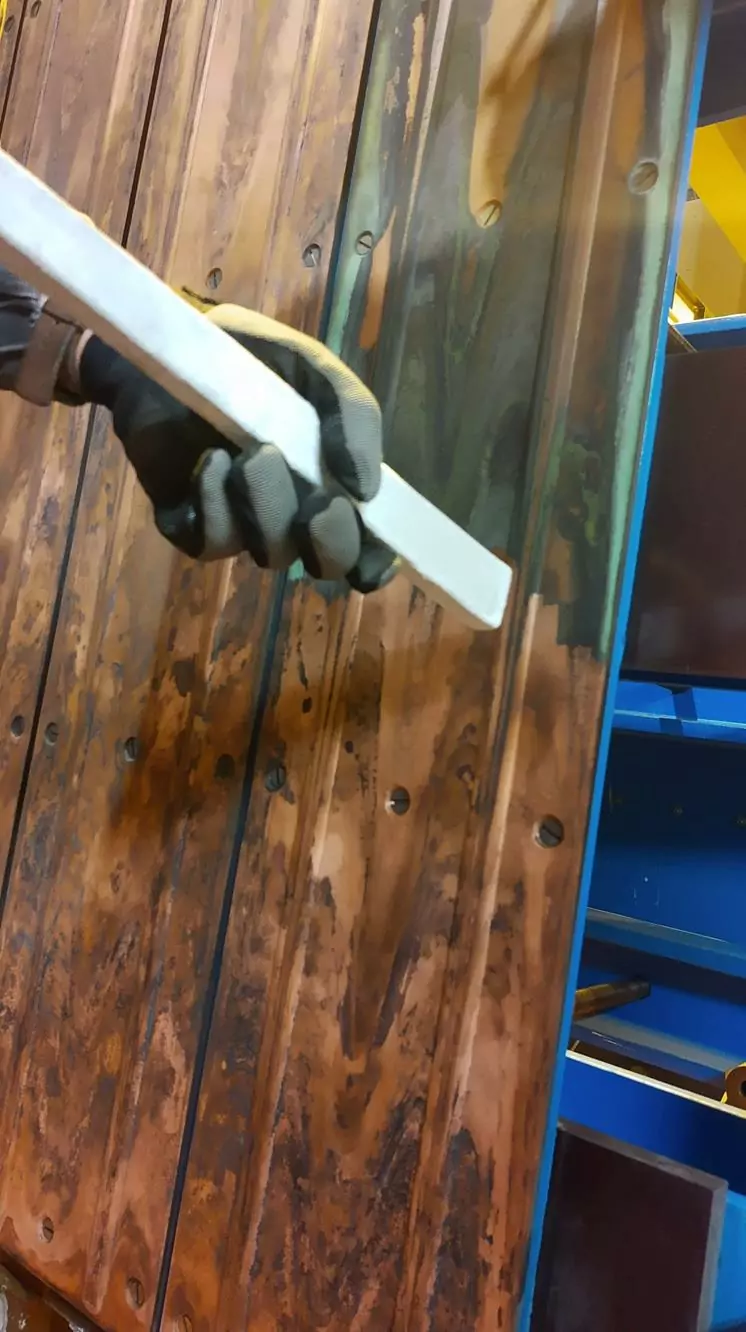 Czyszczenie instalacji i urządzeń elektrycznych 16 Dłoń w rękawiczce używa białego narzędzia do zeskrobania drewnianej ściany, odsłaniając ciemniejsze słoje drewna pod spodem - podobnie jak w Czyszczenie Rozdzielni Elektrycznych Suchym Lodem. W tle widoczne są niebieskie półki.